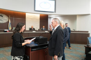 An older man is being sworn in at a ceremony, raising his right hand A woman stands in front of him holding a paper Other individuals are seated at a desk in the background The room has a modern design and a large screen above