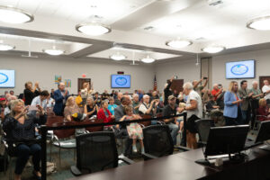 A large group of people, including adults and children, gathered in a well lit conference room Some are standing and some are seated, and several are taking photos with their phones Monitors display a logo The atmosphere appears lively and engaging