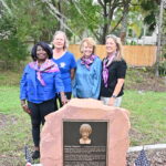 Four women standing in a park beside a memorial plaque on a stone pedestal Two American flags are placed in front The women are smiling and wearing purple scarves, and trees are in the background