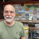 A man with glasses and a beard is smiling while standing in front of a wall covered with license plates He is wearing a green t shirt, and the background includes bar stools and colorful decor