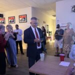 A group of people are gathered in a room with framed pictures on the walls A man in a suit is looking at his phone near a table with drinks Others are interacting and appear to be at a casual social event