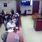 A courtroom scene showing a defendant in an orange jumpsuit standing next to an attorney Several officers and court personnel are present Multiple computer screens are visible at desks, and a large monitor is mounted on the wall