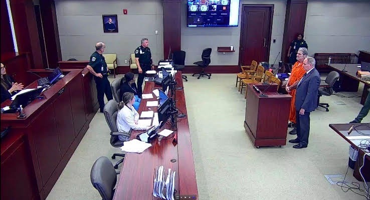 A courtroom scene showing a defendant in an orange jumpsuit standing next to an attorney Several officers and court personnel are present Multiple computer screens are visible at desks, and a large monitor is mounted on the wall