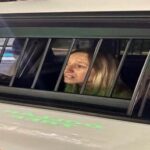 A person with light colored hair is sitting inside a vehicle, looking out through the barred window The interior appears dimly lit, and the person seems to be in a thoughtful or concerned mood