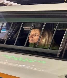 A person with light colored hair is sitting inside a vehicle, looking out through the barred window The interior appears dimly lit, and the person seems to be in a thoughtful or concerned mood