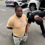 A person is kneeling on the ground with hands behind their back, wearing a beige t shirt and light shorts A police officer is beside them, checking their pockets Police vehicles are parked in the background