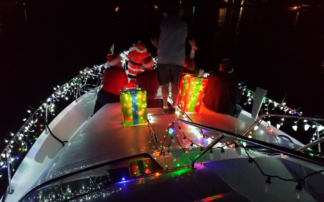 Group of people on a boat decorated with colorful Christmas lights Two large gift props are on the deck, and a person is dressed as Santa The background is a dark, calm water setting with subtle reflections of lights