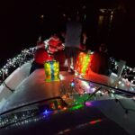 Group of people on a boat decorated with colorful Christmas lights Two large gift props are on the deck, and a person is dressed as Santa The background is a dark, calm water setting with subtle reflections of lights