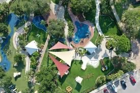 Aerial view of a lush park with a playground, pathways, and a small pool The scene features geometric shade structures, trees, grassy areas, and parked cars at the bottom right