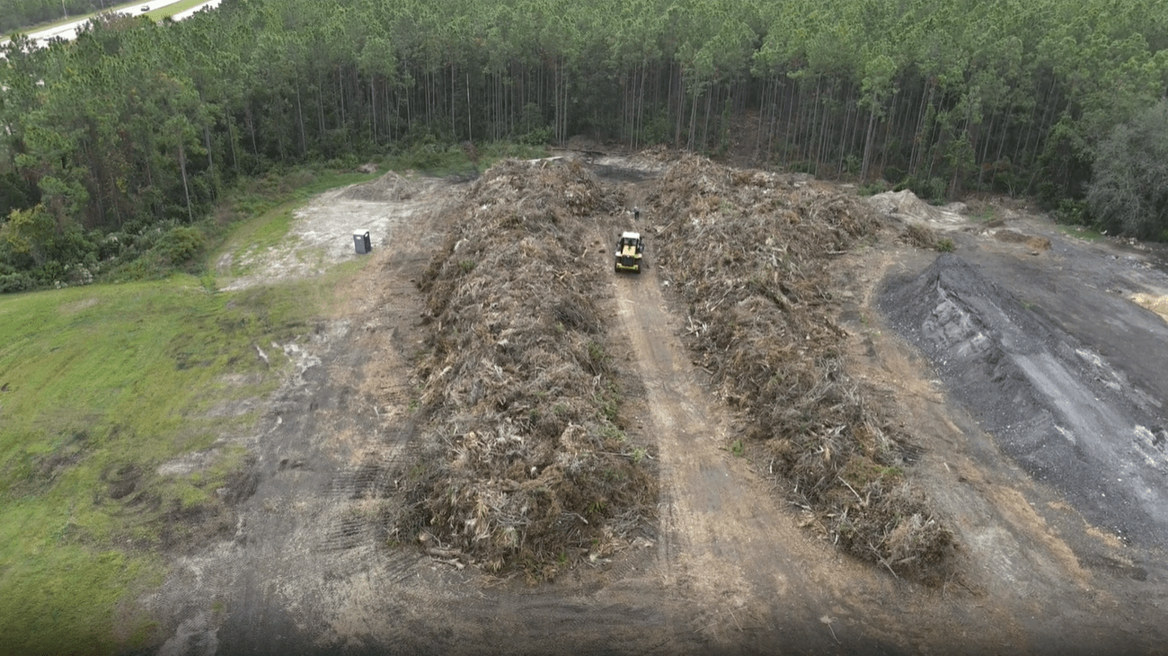 Aerial view of a construction vehicle driving through large piles of tree debris in a clearing near a dense forest Surrounding areas show cleared land and vegetation