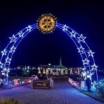 An illuminated arch with glowing stars leads to a festive area at night A star shaped light decorates the top of the arch Trees adorned with lights are visible in the background, along with a sign reading "Rotary"