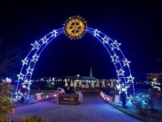 An illuminated arch with glowing stars leads to a festive area at night A star shaped light decorates the top of the arch Trees adorned with lights are visible in the background, along with a sign reading "Rotary"