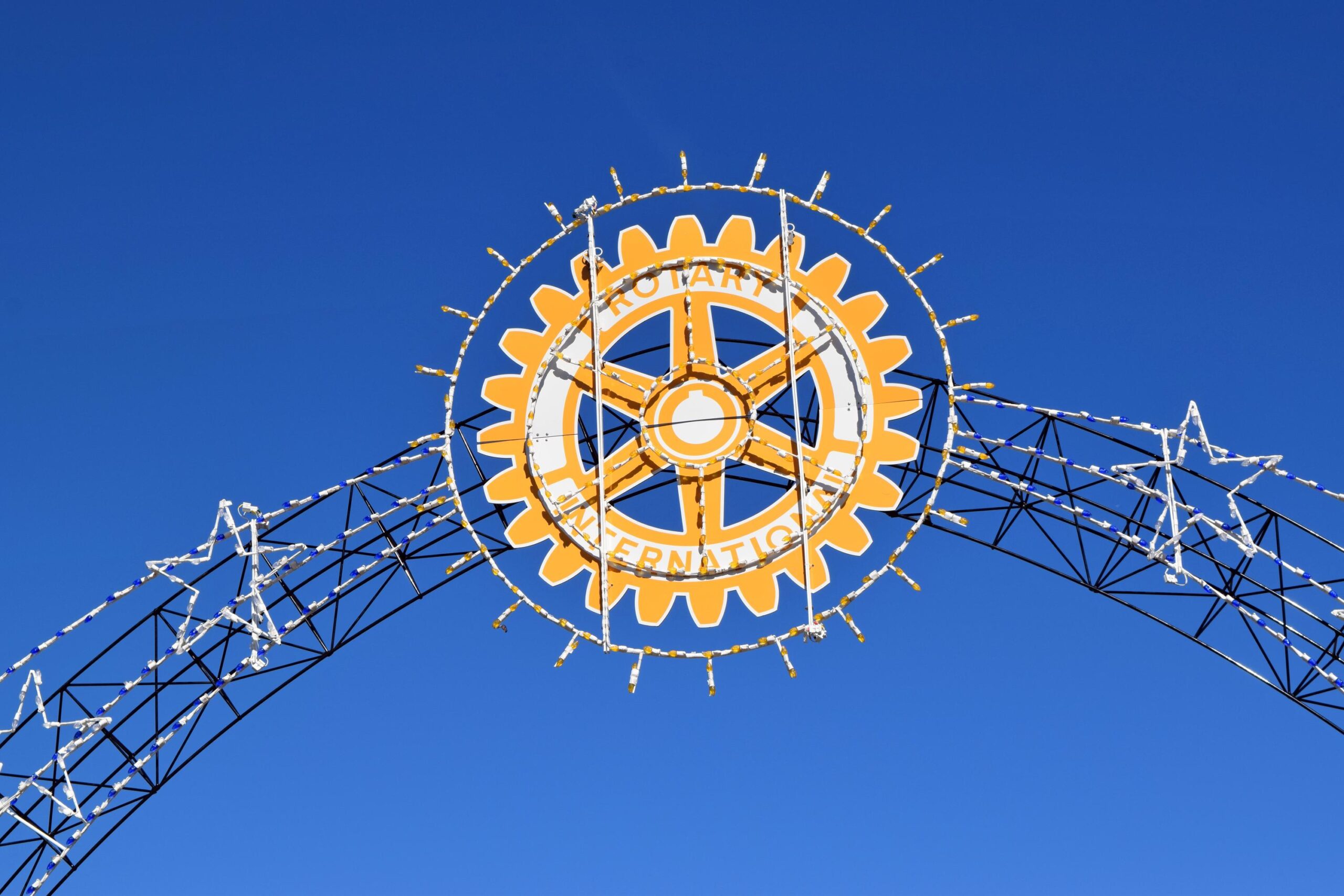 A large, illuminated Rotary International symbol with gear design and white stars is displayed on a metal arch against a clear blue sky