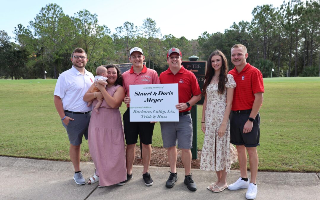 A group of seven people standing outdoors on a golf course, holding a large sign Some are casually dressed, while others wear golf attire Trees and grass are visible in the background, creating a peaceful, green setting