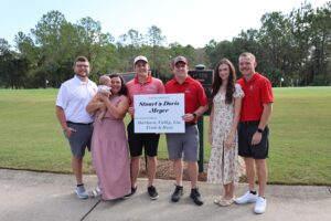 A group of seven people standing outdoors on a golf course, holding a large sign Some are casually dressed, while others wear golf attire Trees and grass are visible in the background, creating a peaceful, green setting