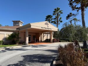 A single story building with a beige facade and covered entrance, labeled "Stuart F Meyer Hospice House " It's surrounded by palm trees and landscaping under a clear blue sky A path leads to the entrance