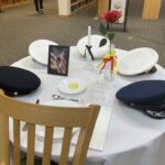 A round table with a white tablecloth holds three military caps, a framed photo, a single red rose, a candle, and an empty plate with a lemon slice There are also scattered confetti stars and an upside down glass