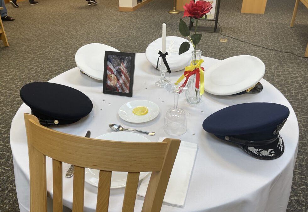 A round table with a white tablecloth holds three military caps, a framed photo, a single red rose, a candle, and an empty plate with a lemon slice There are also scattered confetti stars and an upside down glass