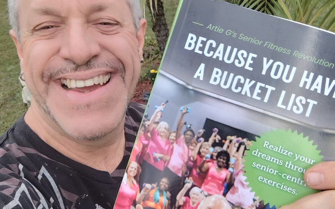 A man smiling and holding a book titled "Because You Have a Bucket List" with a cover image of a happy group of seniors exercising Palm trees and greenery are in the background