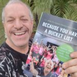 A man smiling and holding a book titled "Because You Have a Bucket List" with a cover image of a happy group of seniors exercising Palm trees and greenery are in the background