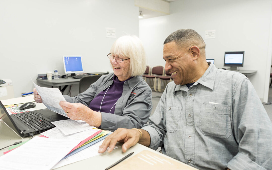 An older woman and a man sit at a table with a laptop and papers They are smiling and appear to be working together in an office setting Computers and chairs are visible in the background