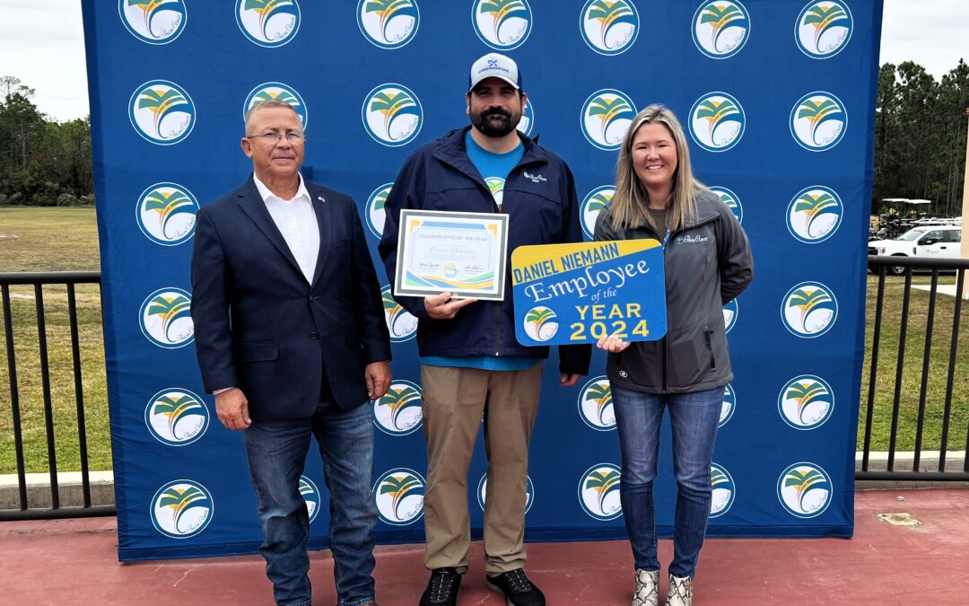 Three people stand in front of a backdrop with a circular design The person in the middle holds a certificate, and the person on the right holds a sign saying "Employee of the Year " They are outdoors on a patio