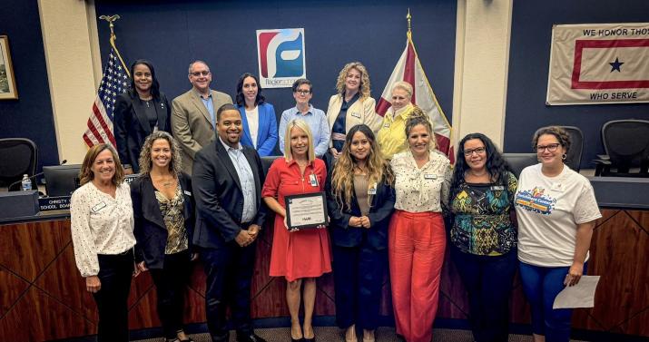 A group of people standing in a room, with one person in the center holding a certificate They are in front of a table and chairs, and there are flags and a banner on the wall behind them