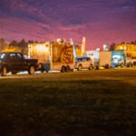 A row of colorful food trucks and parked cars under a vibrant purple and orange evening sky Trees and warm streetlights create a cozy atmosphere at the outdoor event