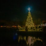 A large Christmas tree adorned with lights stands beside a dark, reflective lake A bright star tops the tree People gather around, with festive lights and tents visible in the background, creating a warm, holiday atmosphere