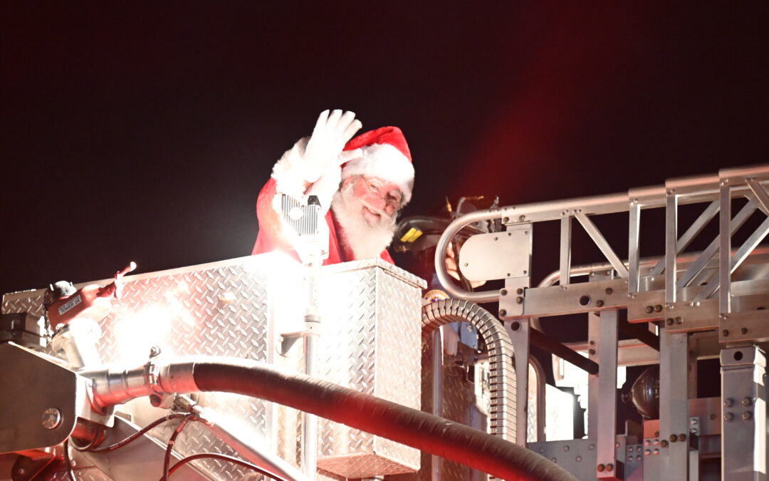 Santa Claus waves from a fire truck's elevated platform at night, surrounded by bright lights and metallic structures