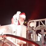 Santa Claus waves from a fire truck's elevated platform at night, surrounded by bright lights and metallic structures