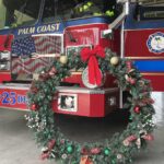 A red fire truck from Palm Coast is parked in a station It has a decorative wreath adorned with a large red bow, ornaments, and ribbons placed in front of it The wreath partially covers the truck's grill and "LAD DE" is visible on the truck