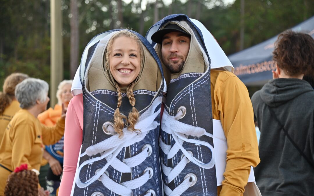 A smiling couple dressed in giant sneaker costumes stand closely together They are outdoors, surrounded by people wearing casual fall clothing Trees and a tent are visible in the background