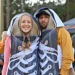 A smiling couple dressed in giant sneaker costumes stand closely together They are outdoors, surrounded by people wearing casual fall clothing Trees and a tent are visible in the background