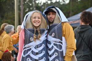 A smiling couple dressed in giant sneaker costumes stand closely together They are outdoors, surrounded by people wearing casual fall clothing Trees and a tent are visible in the background