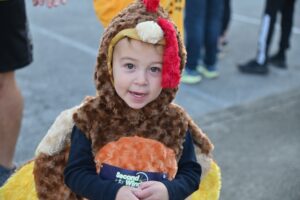 A small child wearing a fuzzy chicken costume stands outdoors The costume includes a red comb and a yellow beak The child smiles at the camera Other people in casual clothing are visible in the blurred background