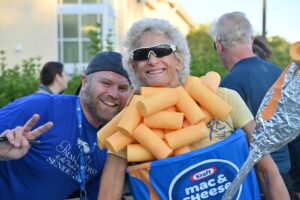 A smiling person in a blue shirt and cap flashes a peace sign, standing next to another person wearing sunglasses and a costume resembling a macaroni and cheese box with yellow foam noodles They are outdoors with others in the background