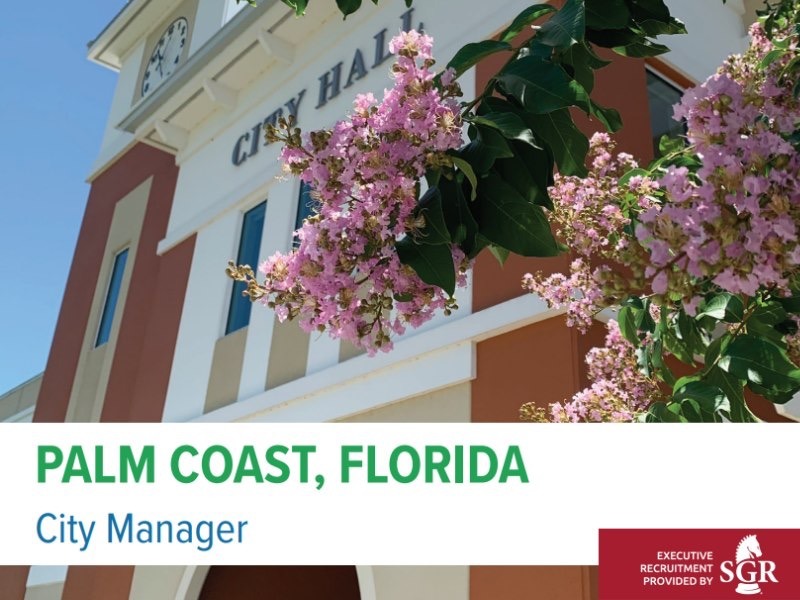 City Hall building in Palm Coast, Florida, featuring blooming pink flowers in the foreground The image announces a City Manager position with a banner displaying "Executive Recruitment Provided by SGR