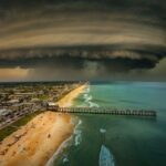 A massive, swirling storm cloud looms over a coastal city, casting shadows over a beach and pier The turbulent sky contrasts with the calm ocean and buildings, creating a dramatic scene