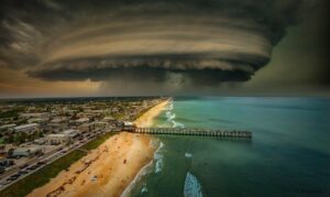 A massive, swirling storm cloud looms over a coastal city, casting shadows over a beach and pier The turbulent sky contrasts with the calm ocean and buildings, creating a dramatic scene