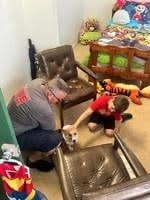 A man and a child are in a room with two chairs and a bed The man is holding a small dog, and the child is reaching out to pet it The bed is covered with colorful bedding and there are toys on the floor