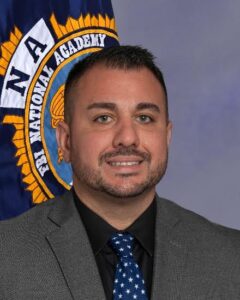 A man wearing a gray suit, black shirt, and blue tie with white stars stands in front of a flag with the words "FBI National Academy