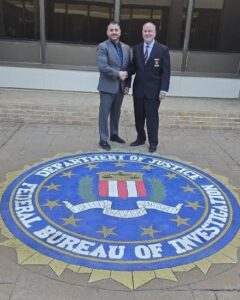 Two people in suits shake hands while standing on a large Federal Bureau of Investigation (FBI) emblem on the ground They are outdoors in front of a building with large windows