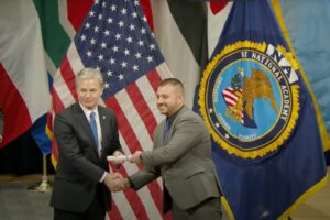 Two men shake hands while exchanging a document in front of the United States flag and the National Academy emblem, surrounded by multiple international flags Both are smiling and dressed in formal attire