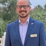 A bald man with glasses and a beard is smiling, wearing a blue suit with a checked shirt and a name tag that reads "Woody " He stands outside with lush greenery and a building in the background