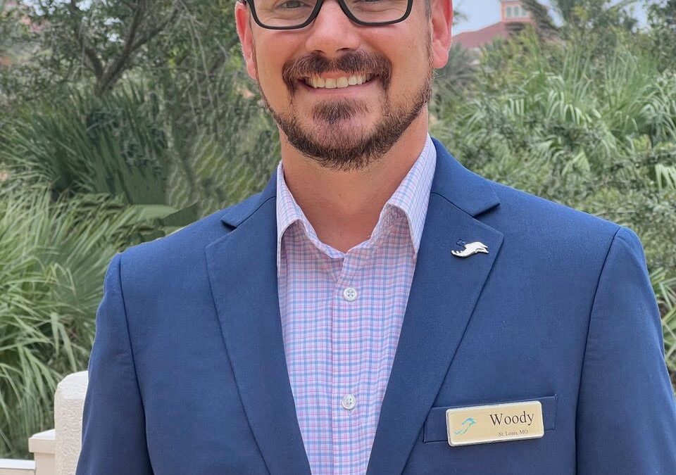 A bald man with glasses and a beard is smiling, wearing a blue suit with a checked shirt and a name tag that reads "Woody " He stands outside with lush greenery and a building in the background