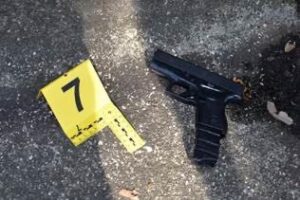 A black handgun lies on the ground next to a yellow evidence marker labeled with the number The surface has a rough, textured appearance with scattered debris