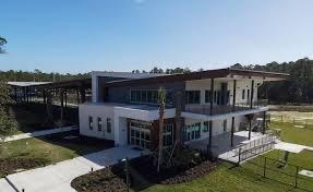 A modern two story building with large windows and a flat roof, surrounded by green lawns and trees The structure features white walls and dark accents, with a covered walkway extending from one side The sky is clear and blue