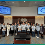 A group of people stands in a council chamber, with some wearing matching shirts A curved desk and a logo reading "Palm Coast" are visible in the background The room has a modern design with large screens and a textured wall