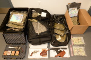 Image of a seizure showing bags of what appears to be marijuana in suitcases and a box, alongside currency in plastic bags, guns, and ammunition boxes